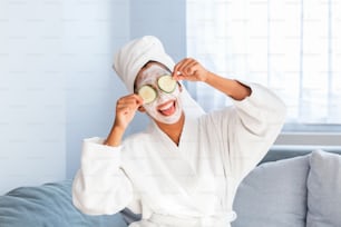 Beautiful young woman with facial mask on her face holding slices of fresh cucumber. Skin care and treatment, spa, natural beauty and cosmetology concept