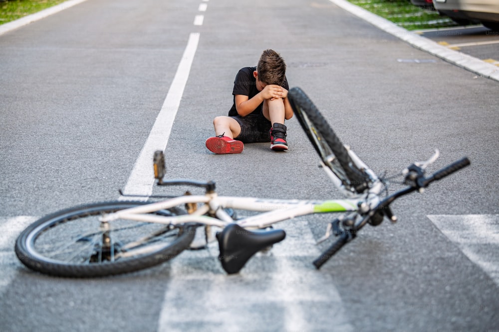 El niño se lastima la pierna después de caerse de la bicicleta. El niño está aprendiendo a andar en bicicleta. Niño en el suelo de la calle con una lesión en la rodilla gritando después de caerse de su bicicleta.