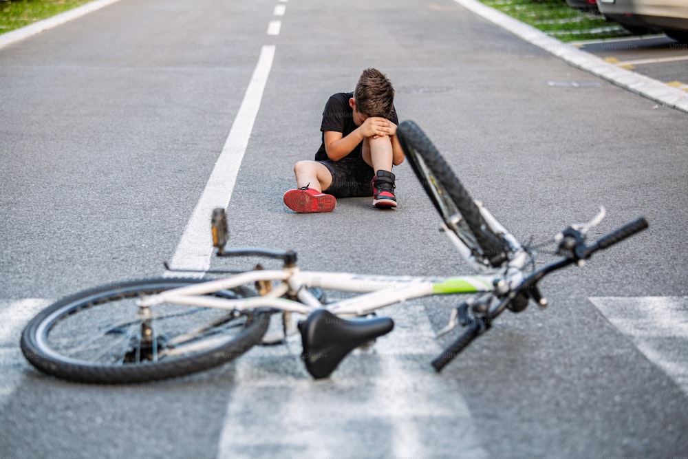 Kid hurts his leg after falling off his bicycle. Child is learning to ride a bike. Boy in the street ground with a knee injury screaming after falling off to his bicycle.