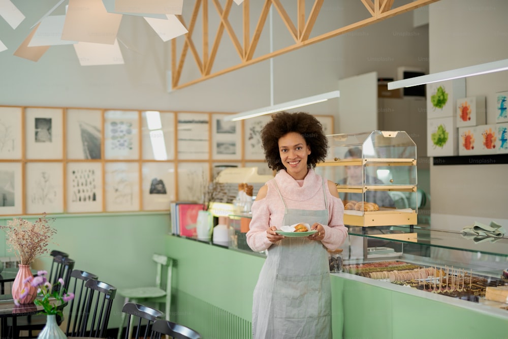Smiling mixed race pastry shop worker holding plate with pastry.