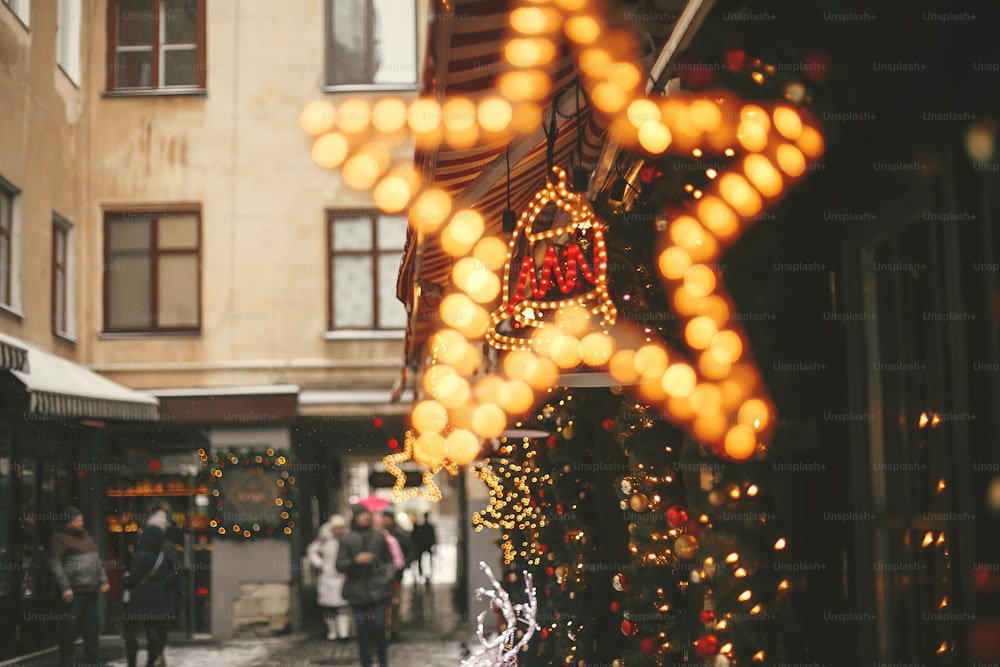 Christmas street decor. Stylish christmas golden star illumination, jingle bell, fir branches with golden lights bokeh on front of building at holiday market in city street