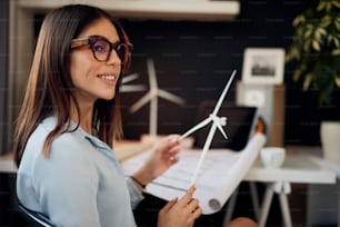 Attractive caucasian businesswoman in suit sitting in modern office and holding windmill model. Sustainable development concept.