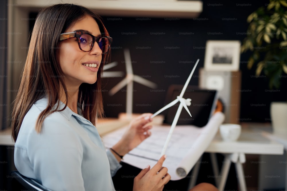Attractive caucasian businesswoman in suit sitting in modern office and holding windmill model. Sustainable development concept.