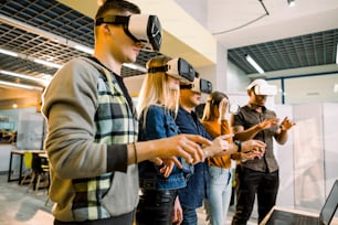 Five multiethnical excited smiling businesspeople wearing virtual reality glasses, interacting with digital interface while standing near table in modern office.