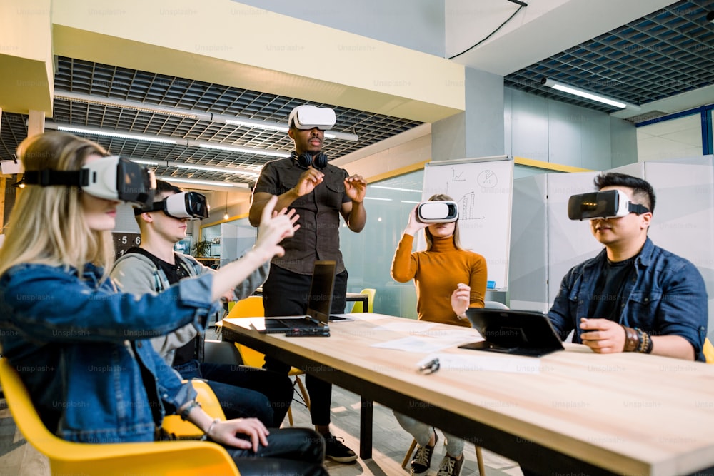 Enjoying new experience. Handsome young African man in VR headset gesturing and smiling while standing at the table. Young people wearing vr goggles.