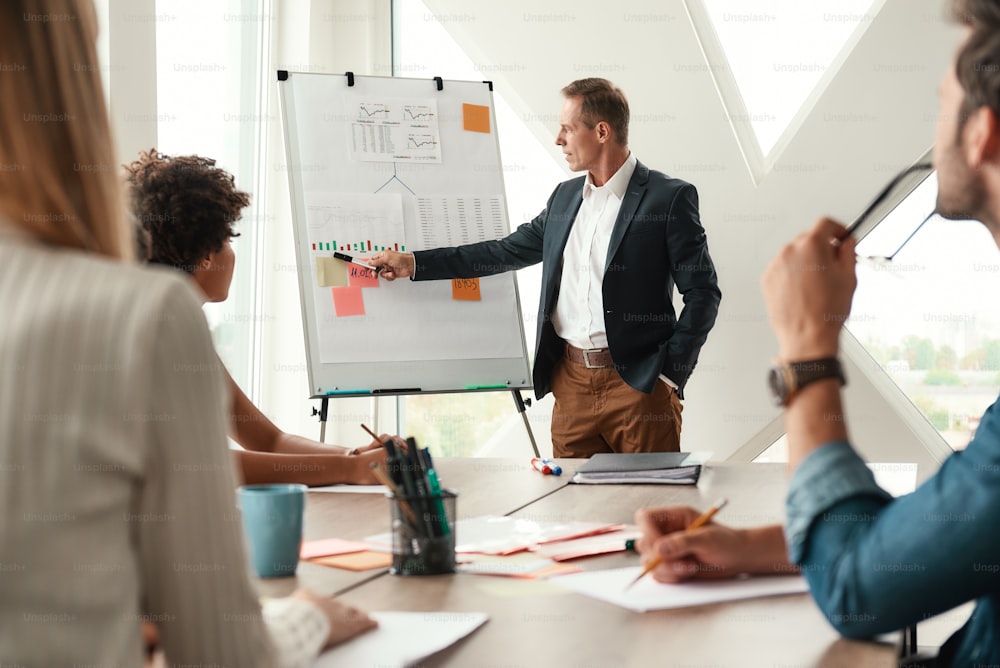 Analyzing results. Mature businessman standing near whiteboard and explaining something to his colleagues. Teamwork. Meeting