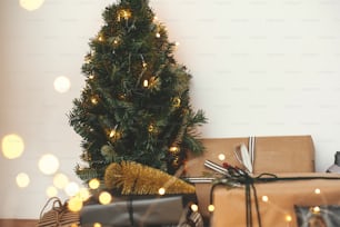 Christmas tree with festive stylish gifts in golden lights bokeh in white room. Happy Holidays. Merry Christmas.Winter holidays preparation. Copy space. Season's Greeting