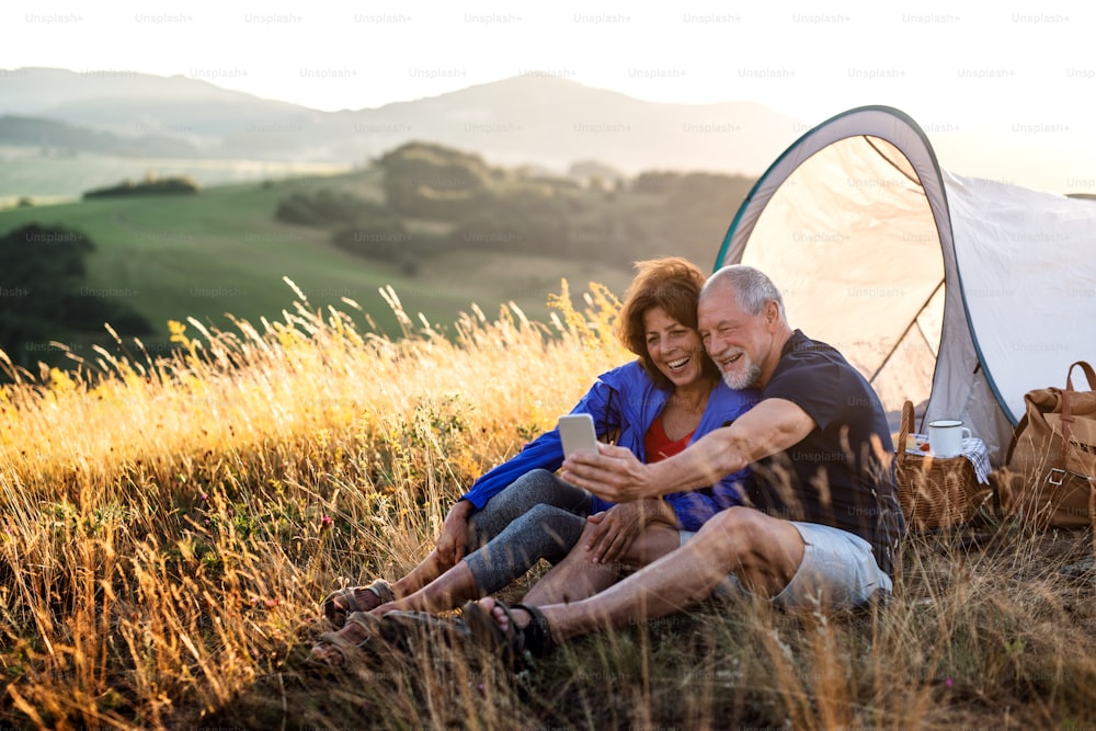A senior tourist couple with smartphone sitting in nature at sunset, taking selfie.
