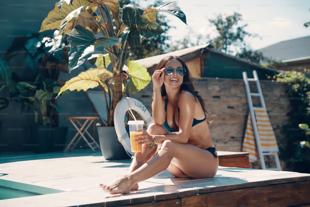 Attractive young woman in bikini drinking cocktail and smiling while sitting by the pool outdoors