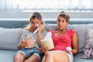 Zwei sentimentale Freundinnen, die weinen und Tränen mit Taschentüchern abwischen, während sie dramatische, traurige Filme, TV-Reality-Shows oder alte Heimvideos auf dem Laptop berühren, während sie zu Hause auf dem Sofa sitzen