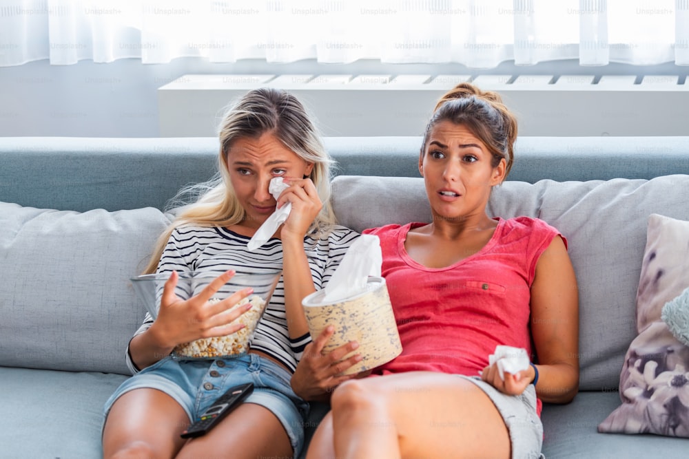 Two sentimental women friends upset crying and wiping tears with handkerchiefs while watching dramatic, sad movie, TV reality show or touching old home video on laptop while sitting on sofa at home