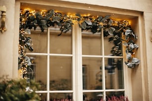 Stylish christmas mistletoe branches with golden  festive lights on window store at holiday market in city street. Christmas street decor. Space for text.