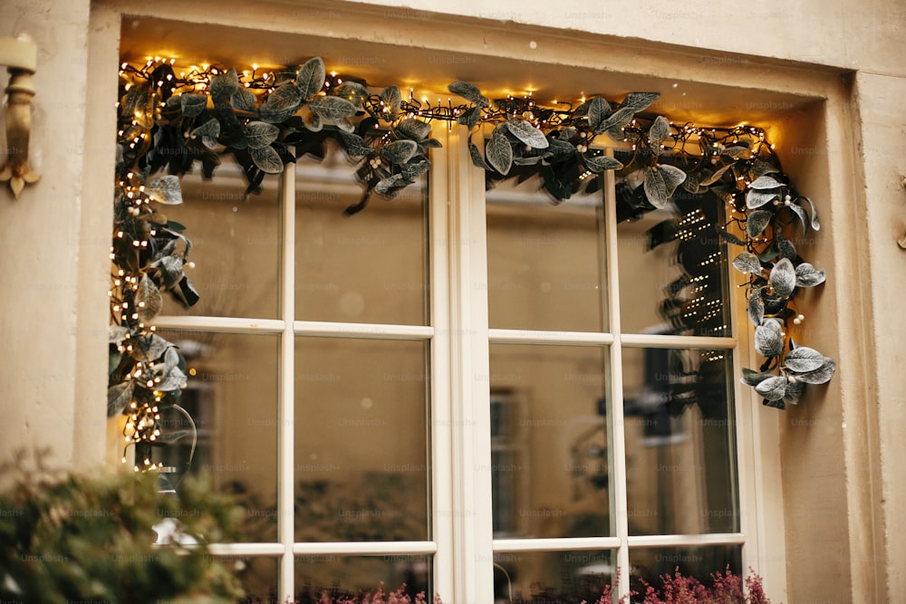 Stylish christmas mistletoe branches with golden  festive lights on window store at holiday market in city street. Christmas street decor. Space for text.