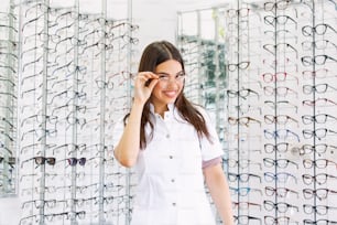 Attractive young female doctor in ophthalmology clinic. Doctor ophthalmologist is standing near shelves with different eyeglasses.