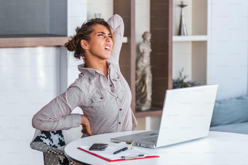 Retrato de una joven mujer estresada sentada en el escritorio de la oficina en casa frente a la computadora portátil, tocando el cuello dolorido con expresión de dolor, sufriendo de dolor de cuello después de trabajar en la PC