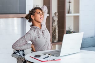 Porträt einer jungen gestressten Frau, die am Home-Office-Schreibtisch vor dem Laptop sitzt, den schmerzenden Nacken mit schmerzerfülltem Ausdruck berührt, nach der Arbeit am PC an Nackenschmerzen leidet