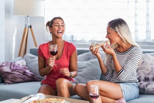 Happy young female friends eating pizza with wine on sofa at home. Happy smiling young women friends eating pizza and opening boottle of wine at home