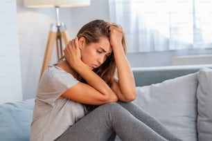 Ritratto di una donna attraente seduta su un divano di casa con mal di testa, dolore e con un'espressione di malessere.