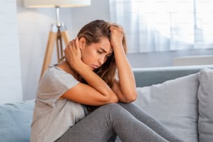 Portrait of an attractive woman sitting on a sofa at home with a headache, feeling pain and with an expression of being unwell.