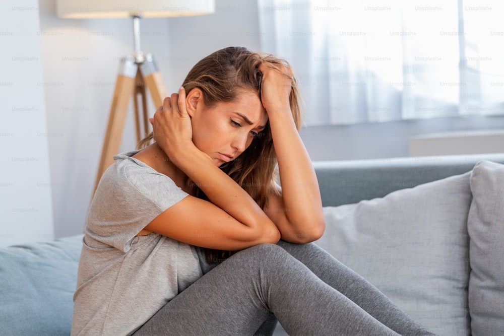Retrato de una mujer atractiva sentada en un sofá de su casa con dolor de cabeza, sintiendo dolor y con una expresión de malestar.