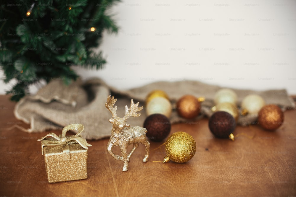 Goldenes Rentier, Geschenkbox und Glitzerkugelspielzeug auf dem Hintergrund des Weihnachtsbaums mit Lichtern auf rustikalem Hintergrund. Frohe Weihnachten und schöne Feiertage. Festliche Dekoration