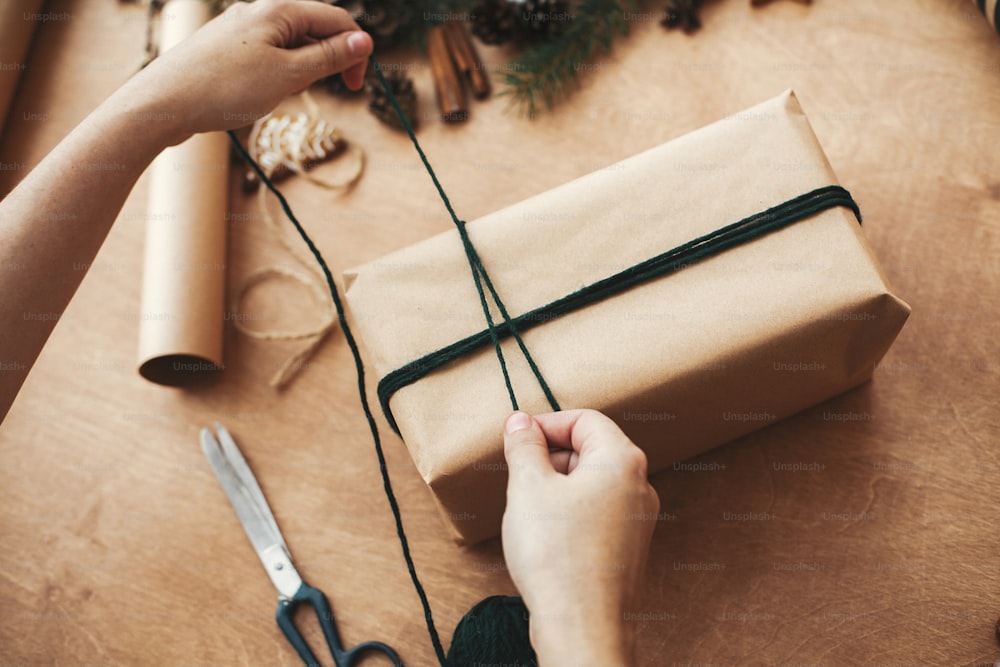 Mains emballant le cadeau de Noël rustique, attachant de la ficelle verte et des branches de pin, des cônes, des biscuits en pain d’épice, du fil, de la cannelle sur une table en bois rurale. Emballage élégant concept de cadeaux de Noël