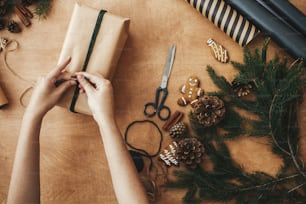 Wrapping stylish christmas gifts. Hands wrapping rustic christmas gift, tying green twine ,and pine branches, cones, gingerbread cookies, thread, cinnamon on rural wooden table.
