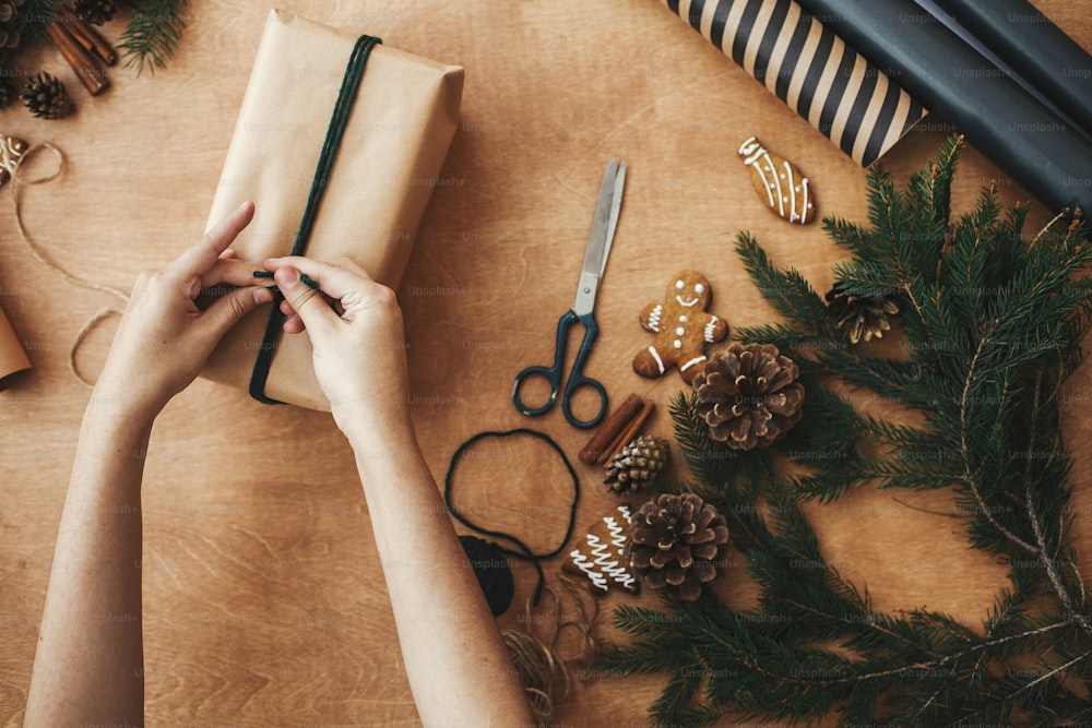 Impacchettare eleganti regali di Natale. Mani che avvolgono il regalo di Natale rustico, legando spago verde e rami di pino, coni, biscotti di pan di zenzero, filo, cannella su un tavolo di legno rurale.
