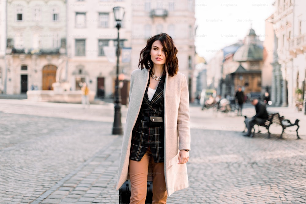 Hübsche junge Frau mit kurzen dunklen Haaren in lässiger stilvoller Kleidung, die mit einem Rollkoffer reist und auf der Straße der alten europäischen Stadt spazieren geht.