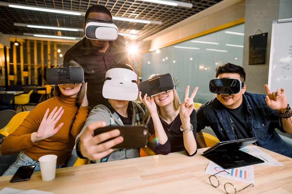 Five young funny people sitting at the table in front of each other, using virtual reality goggles, having fun and gesturing. VR goggles concept, business meeting