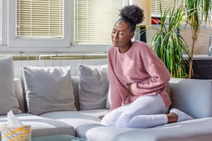 Jeune femme noire souffrant de maux de ventre sur le canapé à la maison. Femme assise sur un lit et ayant mal au ventre. Jeune femme souffrant de douleurs abdominales assise sur un canapé à la maison