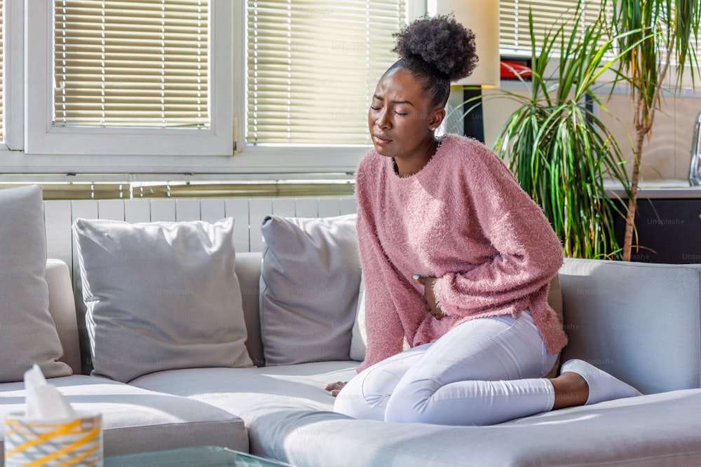 Young black woman suffering from stomachache on sofa at home. Woman sitting on bed and having stomach ache. Young woman suffering from abdominal pain while sitting on sofa at home