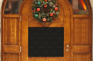 Stylish christmas wreath with red and green baubles and empty board with copy space on rustic wooden doors at holiday market in city street. Christmas template. Space for text
