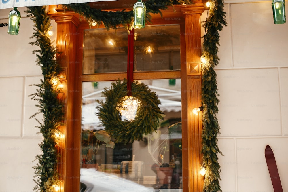 Stylish christmas wreath with fir branches and lights at front window of store at holiday market in city street. Space for text. Rustic decoration. Christmas street decor.