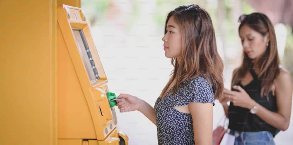 Duas jovens mulheres asiáticas retirando o dinheiro de um cartão bancário usando caixa eletrônico no shopping
