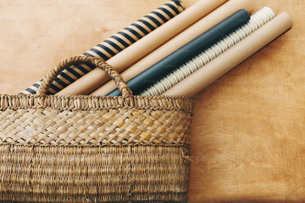 Stylish christmas wrapping paper rolls in rustic straw basket on wooden table, copy space. Preparation for winter holidays. Craft, black and gold wrapping papers