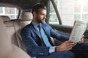Latest news. Young confident businessman in formal wear reading fresh newspaper while sitting on the back seat of his car. Media concept. Business trip. Success concept
