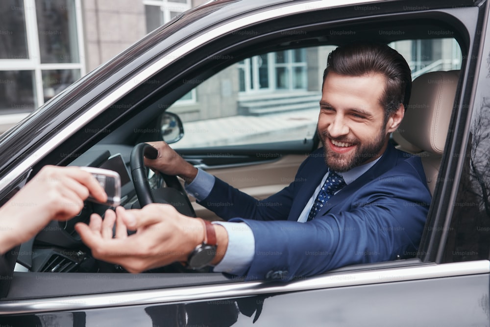 Feliz y exitoso. Un joven hombre de negocios alegre y elegante con traje completo toma una llave del automóvil y sonríe mientras está sentado en el asiento delantero. Emoción positiva. Concepto de negocio. Prueba de manejo