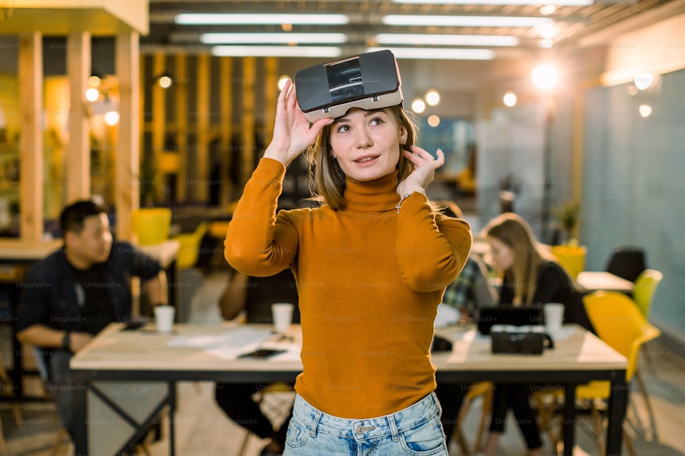 Pretty female business woman in orange sweater trying 3d vr glasses at work. Multiracial team of young people working together in creative office.