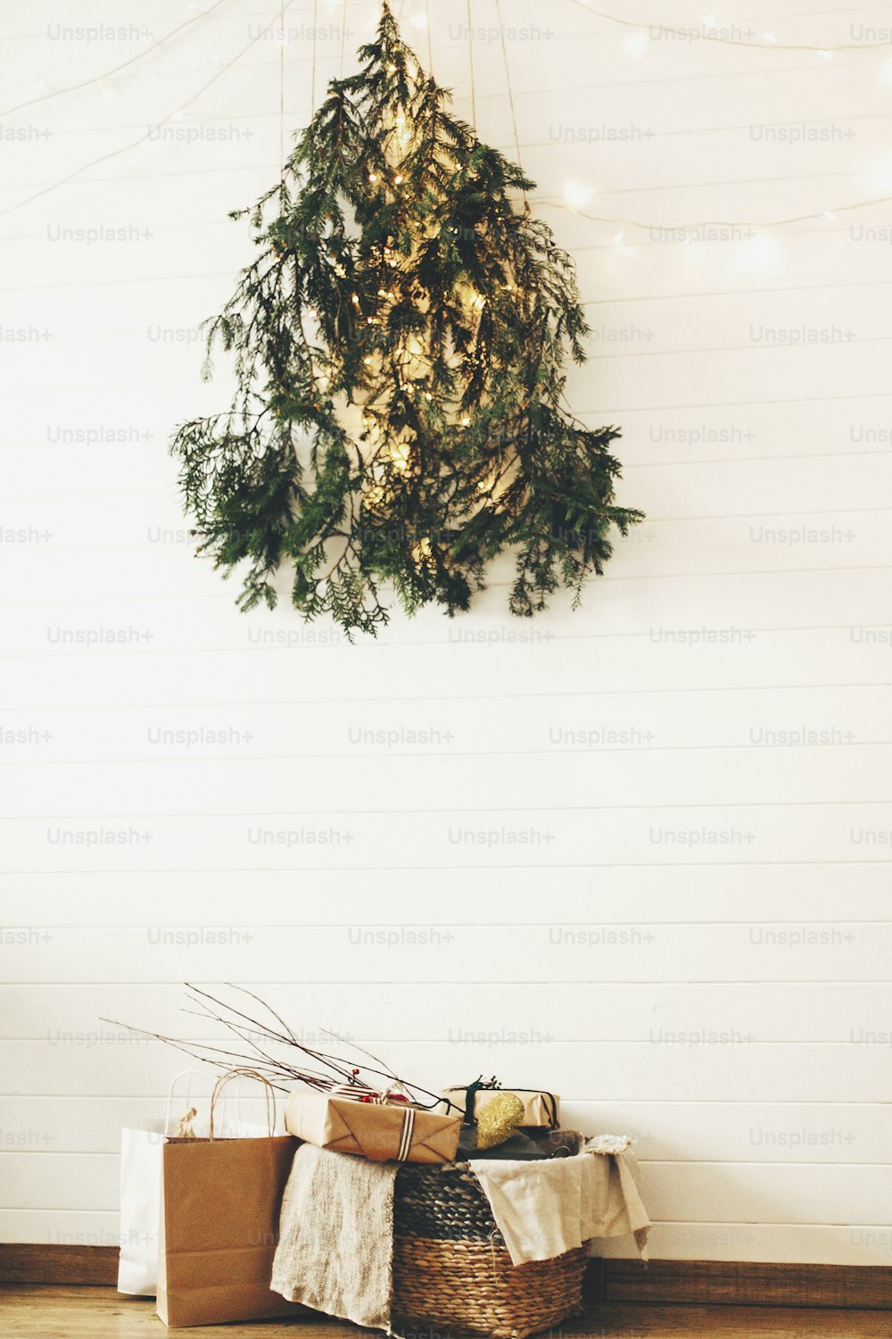Árbol ecológico navideño moderno de ramas de pino que cuelga en la pared con luces festivas y regalos en canasta rústica. Elegantes cajas de regalo de Navidad en piso de madera debajo del árbol de Navidad creativo.