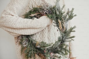 Christmas rustic wreath. Hipster girl in stylish white sweater holding rural christmas wreath with fir branches, berries, pine cones and herbs in room. Happy holidays
