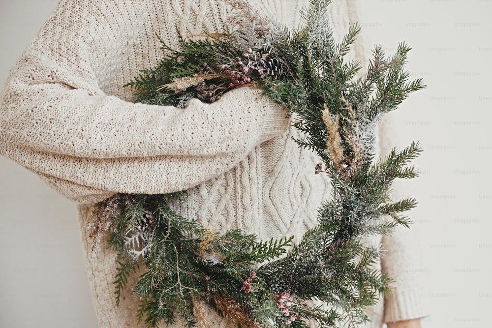 Corona rústica de Navidad. Chica hipster con elegante suéter blanco que sostiene una corona navideña rural con ramas de abeto, bayas, piñas y hierbas en la habitación. Felices Fiestas