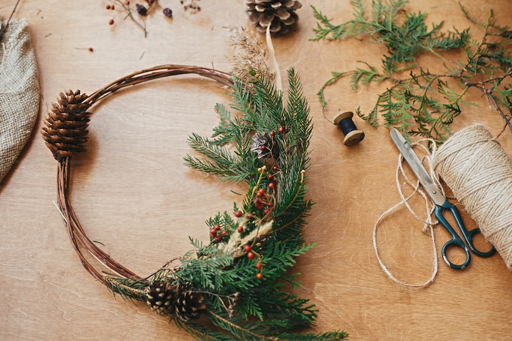 素朴なクリスマスリース。田舎の木製のテーブルに、はさみと糸でモミの枝、ベリー、松ぼっくり、ハーブを使ったクリエイティブなクリスマスリース。楽しい休暇をお過ごしください。工房