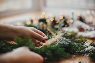 Hands holding herbs and fir branches, pine cones, thread, berries, golden lights on wooden table. Christmas wreath workshop. Authentic stylish still life. Making rustic Christmas wreath.