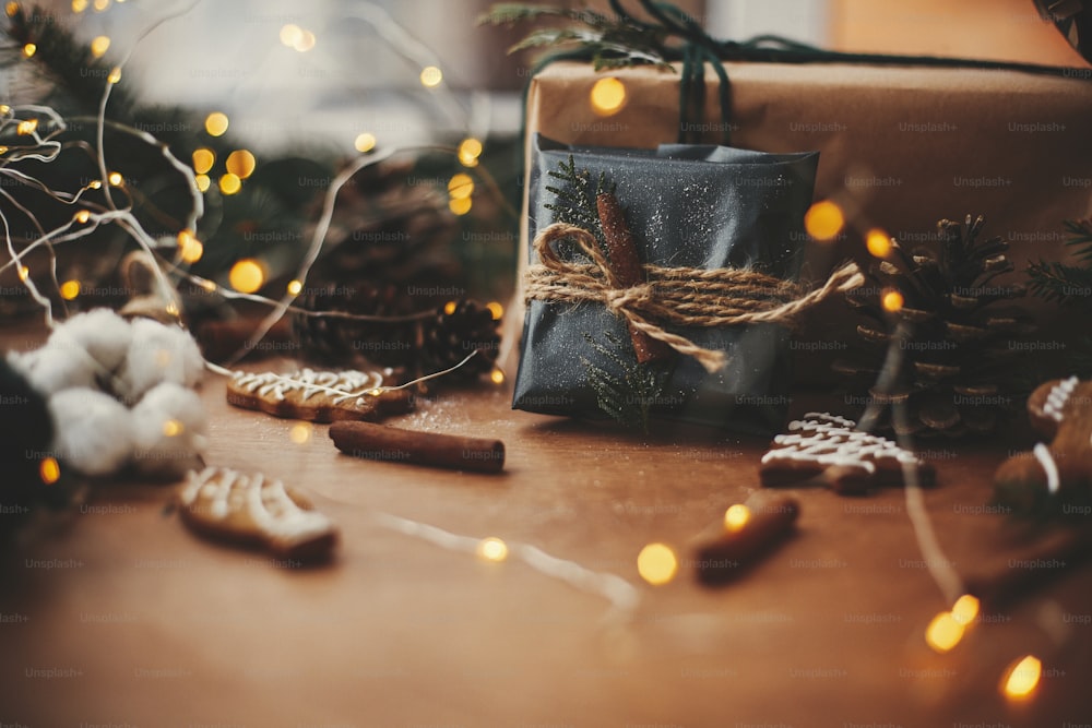 Elegante regalo di Natale rustico in carta da regalo nera con decorazione cannella su sfondo di luci dorate, rami di pino, pigne, biscotti di pan di zenzero e cotone. Buon Natale