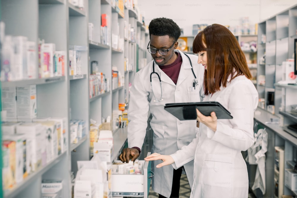 Retrato de dos sonrientes farmacéuticos multiétnicos amigables que trabajan en una farmacia moderna y hacen pedidos de medicamentos en una empresa de distribución.