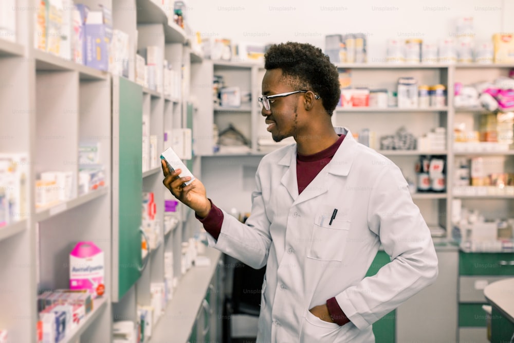 Portrait of African American male pharmacist standing in interior of pharmacy and looking at the medicine in hand.