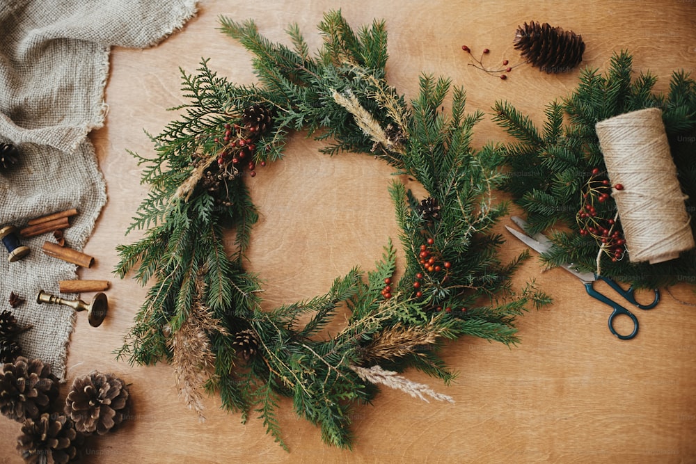 Rustic Christmas wreath flat lay. Fir branches, pine cones, thread, berries, scissors on wooden table. Christmas wreath workshop. Authentic stylish still life. Happy Holidays