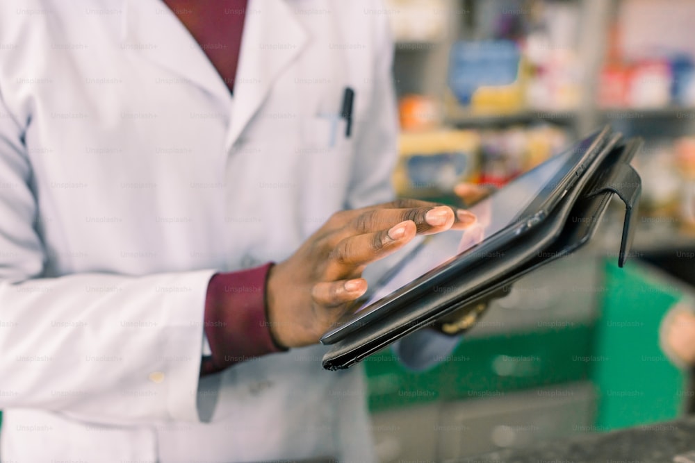Nahaufnahme der Hände eines afroamerikanischen Arztes oder Apothekers mit digitalem Tablet, während er im Inneren einer Apotheke oder eines Krankenhauses steht.
