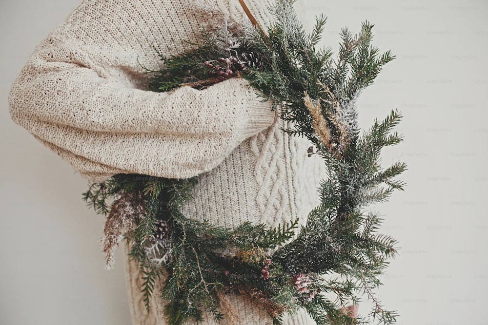 Hipster girl in stylish white sweater holding rural christmas wreath with fir branches, berries, pine cones and herbs in room. Christmas rustic wreath. Season's greetings